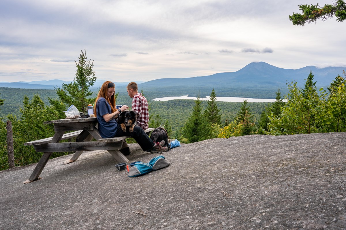 Hikers and a dog sit at a picnic table overlooking Katahdin.