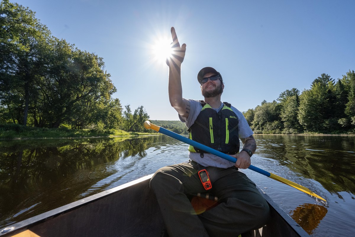 A man sits in the stern of a canoe in a river. He points at something as the sun shines above. 