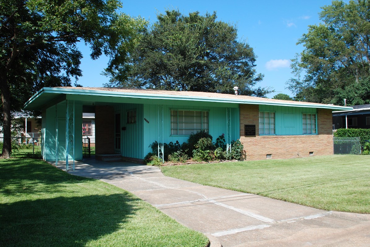 A small ranch-style home, aqua in color with light brown brick accents, sits on a shaded lot. 