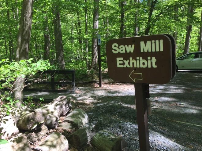 Wooden Sign with the words Saw Mill Exhibit. Behind the sign is a small gravel parking area. 