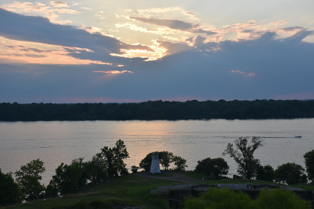 a green shoreline with small lighthouse along river at sunset