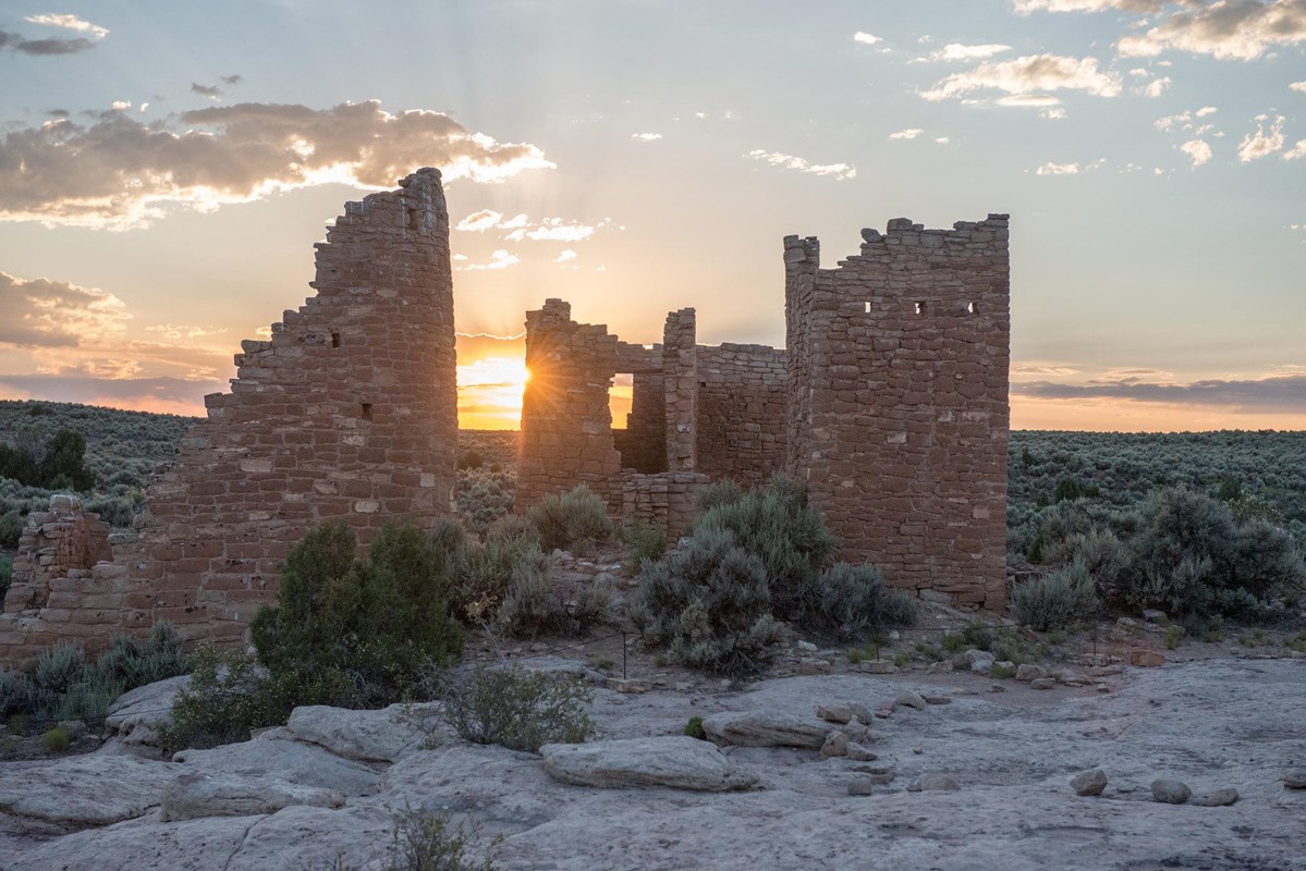 Tall stone structures with a setting sun shining through them. Desert landscape surrounds them.