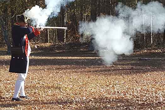 A man is seen holding a musket at his shoulder firing. A plume of smoke is prominent. 