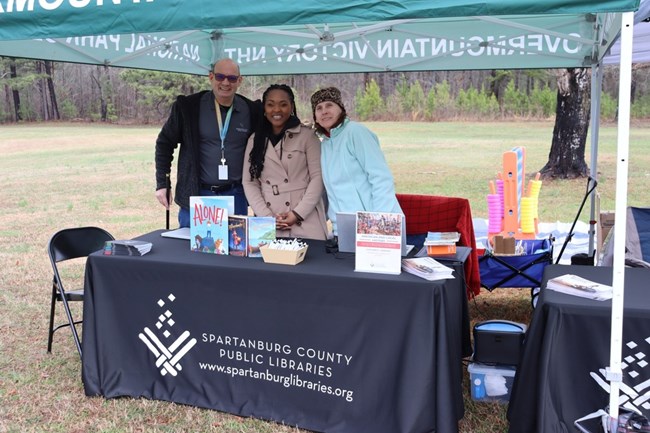 Three libririans stand behind table with blue table cloth under a tent