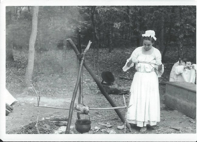 A woman stands near a fire with a chicken roasting over a pot.