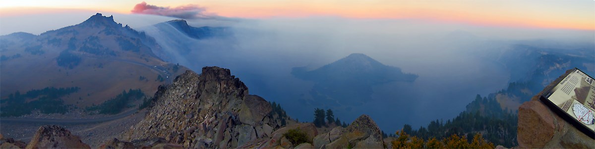 panoramic view from above 8900 feet in elevation of smoke rolling over the caldera rim and obscuring views of the lake and Wizard Island.