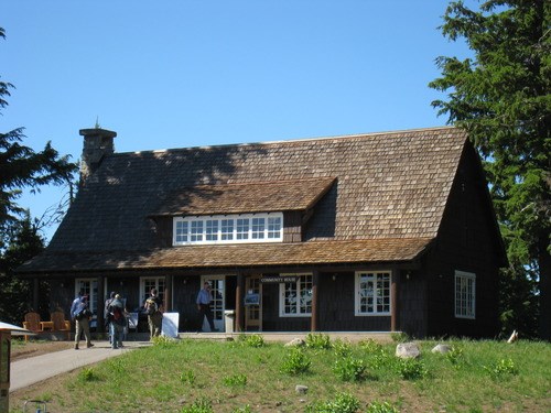 A steeply pitched roof with shaker shingles, and a large dormer window covers a long front porch and a two-story barn-like structure. Visitors walk along the sidewalk.