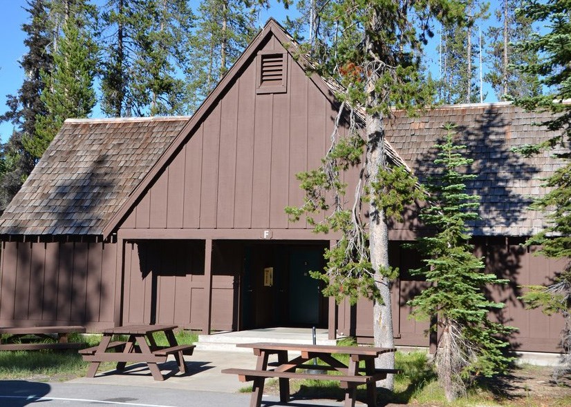 A brown single-story structure with high-pitched roof over a breezeway and entrance doors to accommodations is surrounded by tall lodgepole pines with two picnic table in front.