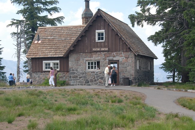 The Rim Visitor Center is an historic building with native stone walls, brown painted upper frame, and wood shake shingle roof.  Several visitors stand outside the front door or walk along the concrete path.