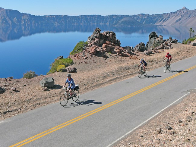Bicyclists riding along East Rim Drive with view of Crater Lake in the background.