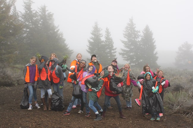 a group of teenagers wearing bright orange vests and raincoats posing for a photo in front of pine trees and shrubs on a foggy day