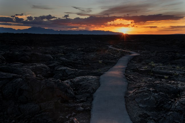 Black asphalt trail meandering over a lava landscape with the sun setting in the distance.