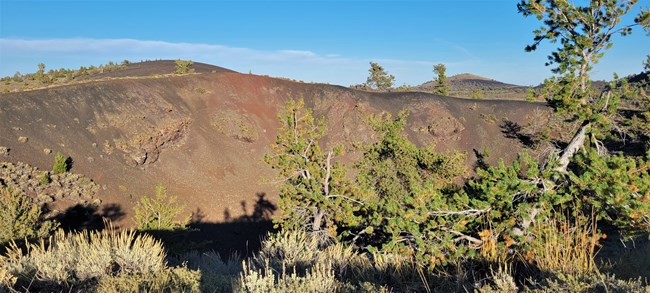 a reddish volcanic crater with sagebrush and pine trees growing along the rim and slopes