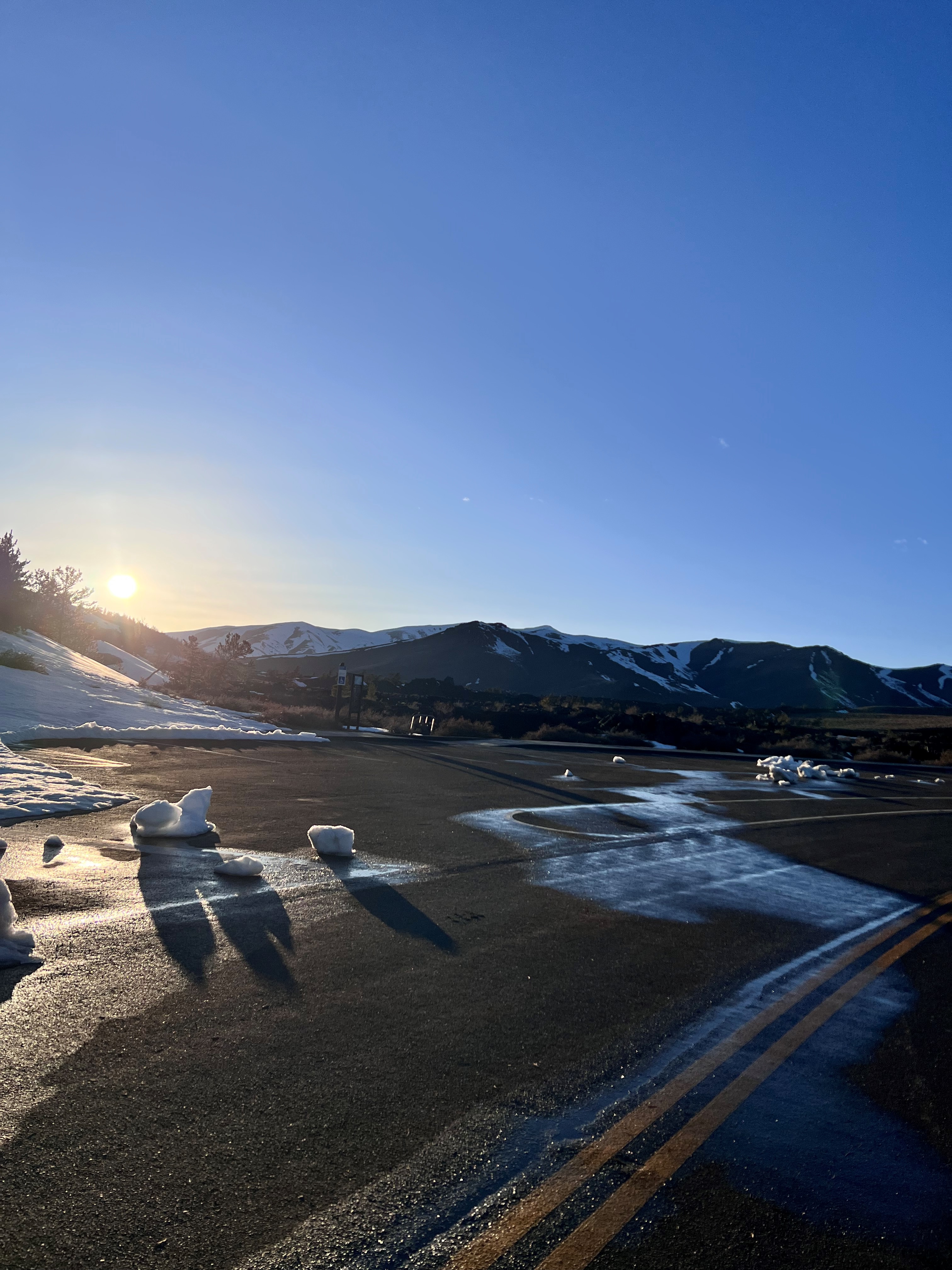 The sun rises over snow capped mountains and illuminates a road and parking lot dotted with melting snow and ice.