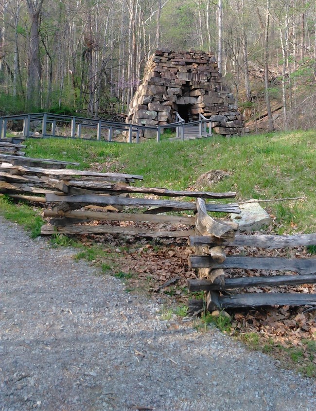 A small stone pyramid shaped structure on top of a hill surrounded by a wooden fence, trees, and grass.