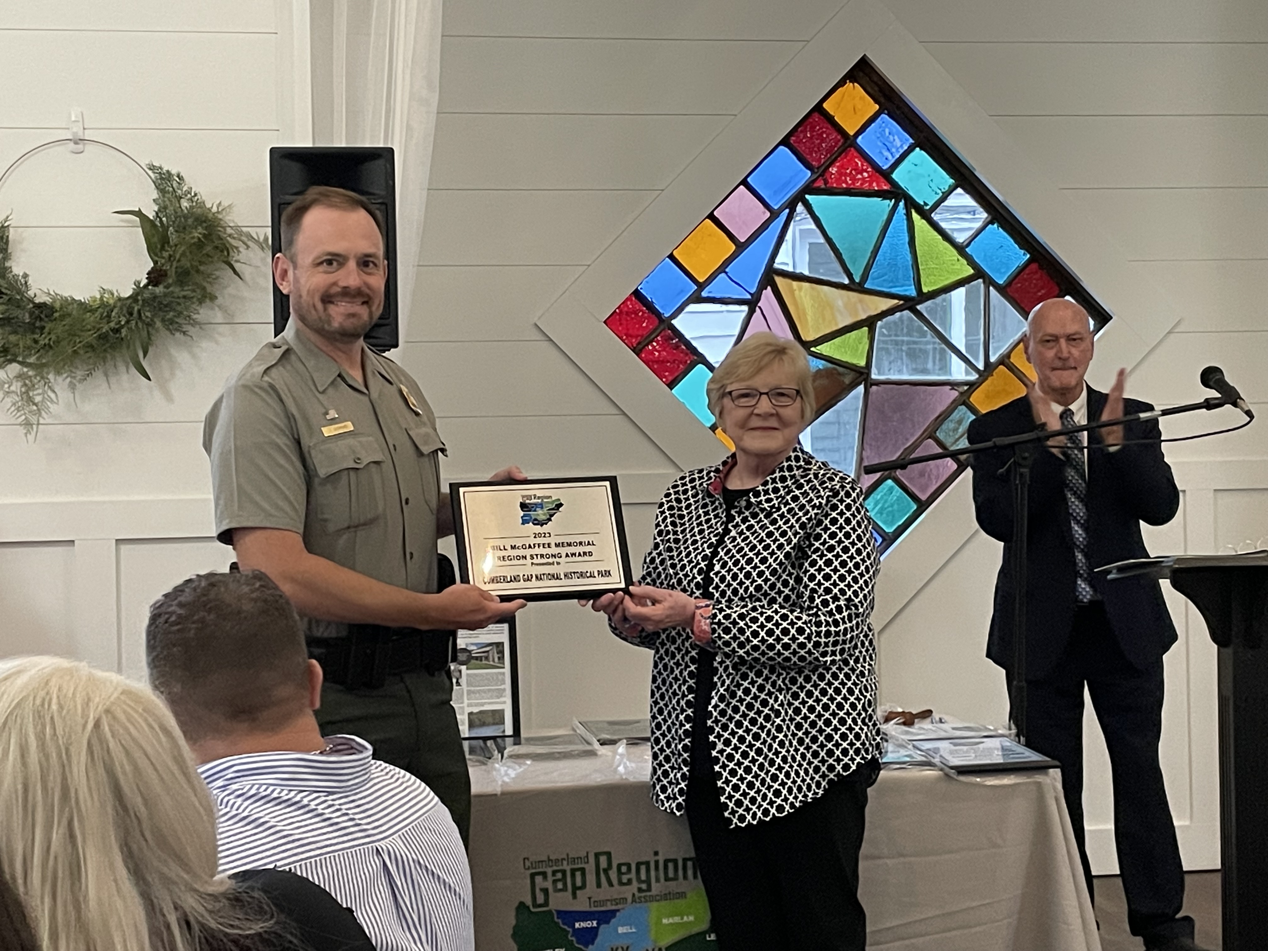 A woman presents a plaque to a man in National Park Service uniform as people applaud.