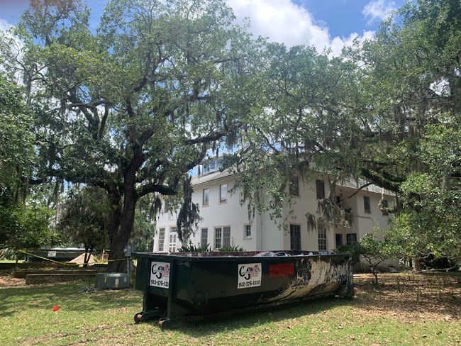 Dumpster sits in front of a two story white building with a pile of dirt surrounded by yellow caution tape and a dumpster in the distance.  Trees surround the house.