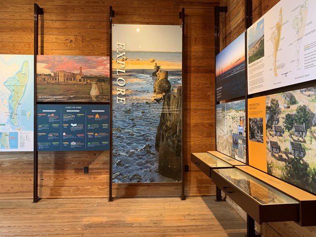View of vertical exhibit panels against a wooden wall showing activities on the island.  The wall next to it has 4 colored panels of natural and cultural history.  Two drawers below the panels open to show artifacts from the island.