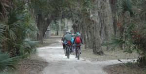 two visitors ride bikes on sandy road under tree canopy