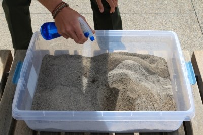 Person holding a spray bottle squirts water into a box of sand