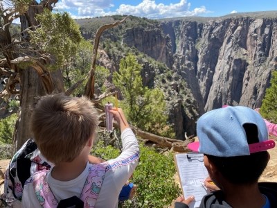 Two children are standing in front of a deep, steep canyon. They are holding a clipboard and thermometer.