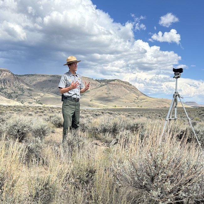 A park ranger wearing a uniform stands in front of a camera on a tripod. Mesas are in the background.