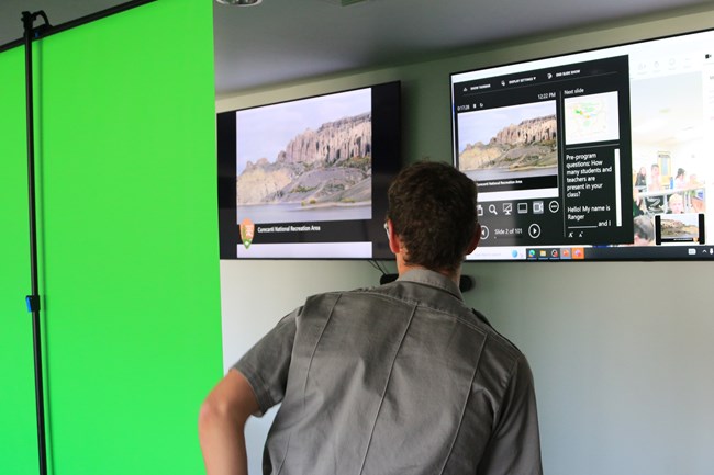 A park ranger wearing a grey shirt stands by a green pop up screen. Two large monitors displaying photos are in the background.