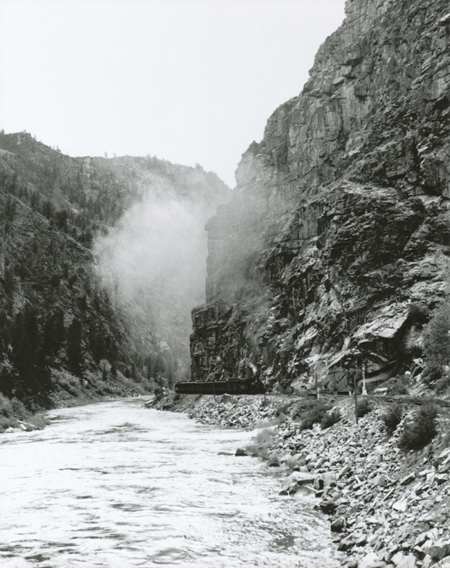 Black and white image of a engine and train cars in a narrow,steep canyon. A river is to the left of the railroad tracks.