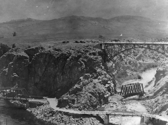 Historic black and white image of four bridges spanning different sections of rivers between steep canyons. Cars are visible next to the highest bridge. Mesas are in the background.