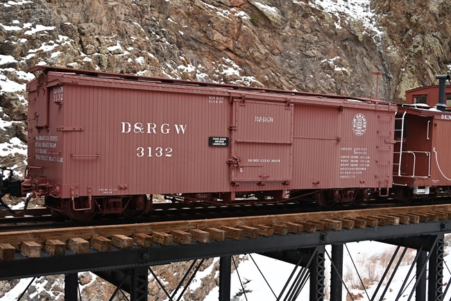 A red boxcar with white lettering sits on a wooden platform. A red caboose is visible in the right corner. Snow and canyon walls are in the background.