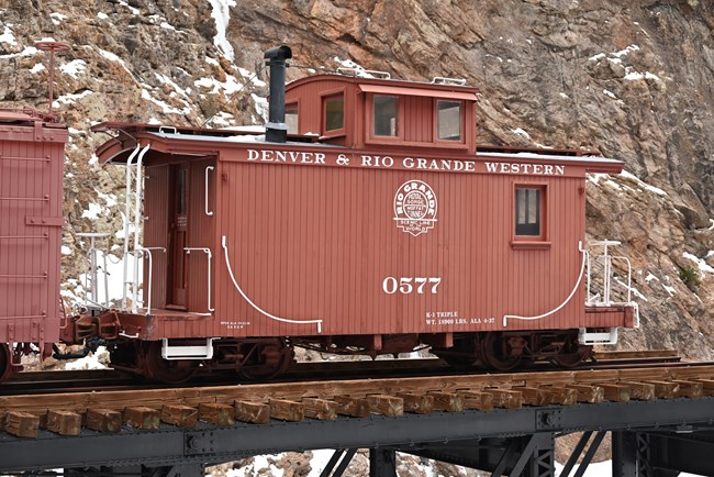 A red caboose railcar sits on a wooden platform atop a steel bridge. Canyon walls and snow are in the background.