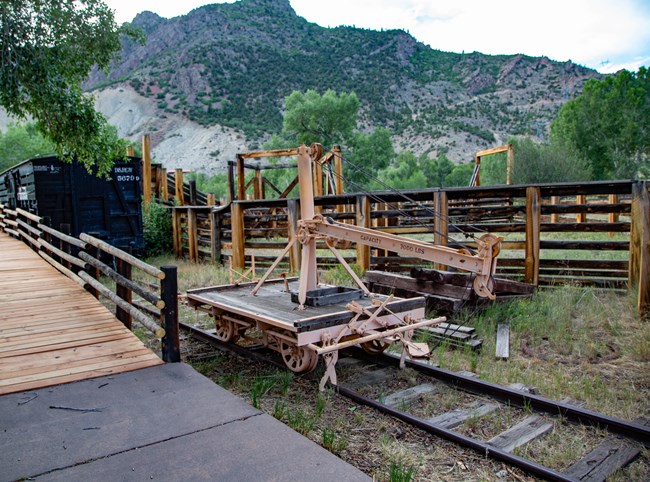 Railroad exhibit with black train car, loading equipment, and wooden corrals. A concrete and wooden walkway is to the left of the exhibit area.