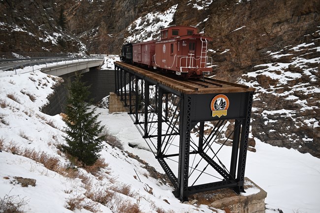 A steel span bridge with a wooden platform has multiple train cars on it. The bridge is in a canyon. Snow covers the ground and surrounding walls of the enbankment.