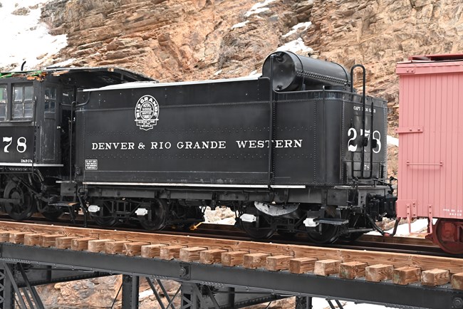 Black coal tender rail car with a black engine on the left and red boxcar on the right. All three cars are on a wooden platform atop a steel bridge.