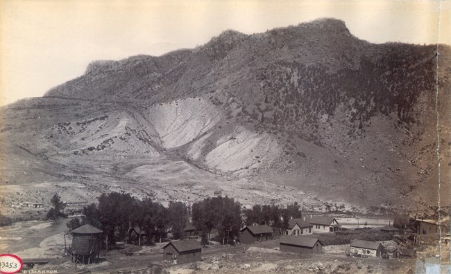 Historic image of a small town with numerous buildings, small water tower, and railroad tracks. A large mesas is behind the town.