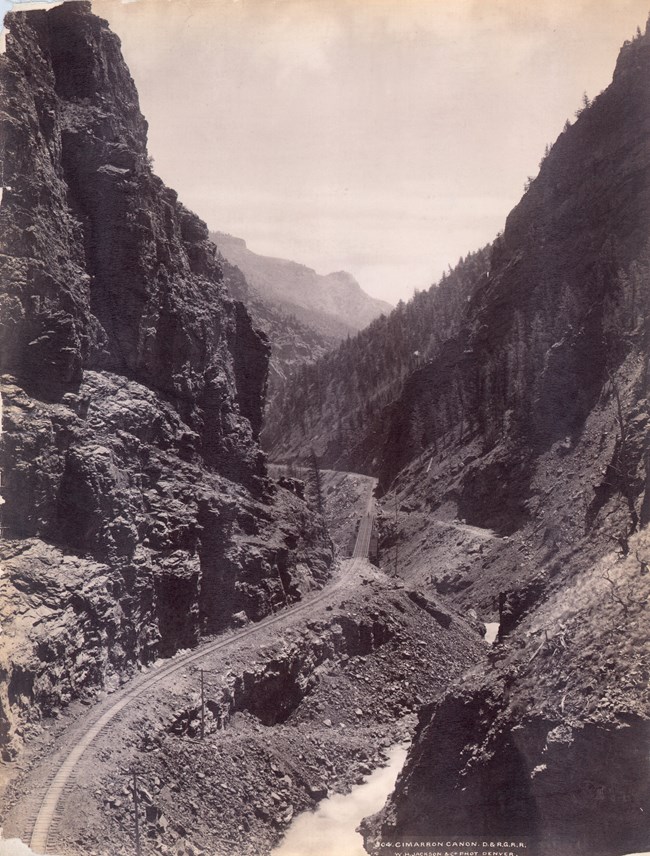 Historic image of a railroad and truss bridge running through a steep, narrow canyon. A river is flowing alongside the railroad. Conifer trees grow on the north facing slopes of the canyon.