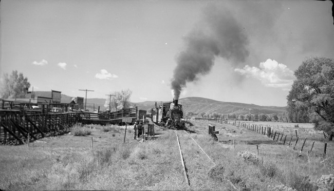 Historic black and white image of a train at a railyard with men, stock pens, a loading chute, and storefronts.