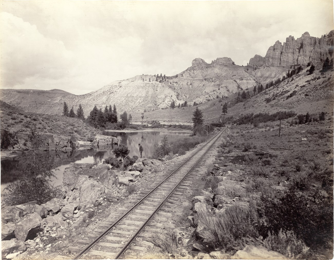 A man stands on a rock by a river. Image shows narrow gauge tracks of the Denver and Rio Grande Railway, telegraph poles, steel rails near the tracks and rock outcroppings in the distance. An excursion train is on the distance.