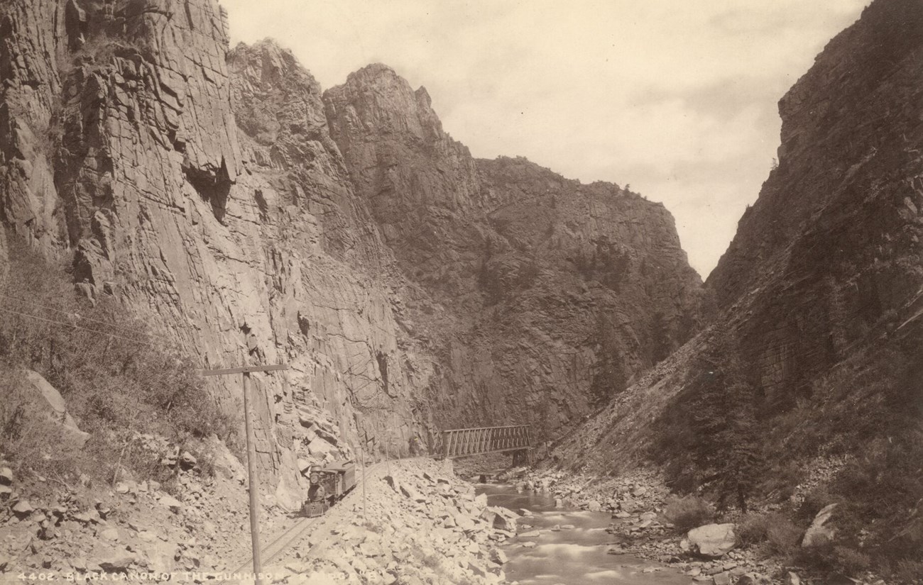 Historic image of an engine rides train tracks laid in a steep, narrow canyon. The railroad runs alongside a river. A small bridge spans the river between canyon walls.