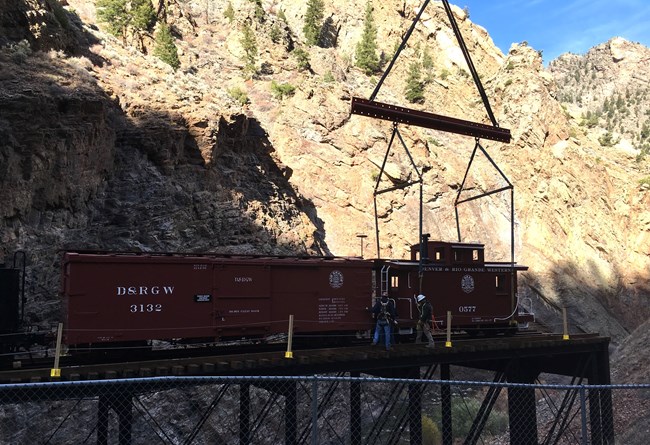 Two dark red train cars are lifted by a crane onto a wood and metal bridge. Tall canyon walls are behind them. Two people wearing harnesses and hard hats stand by the train cars.