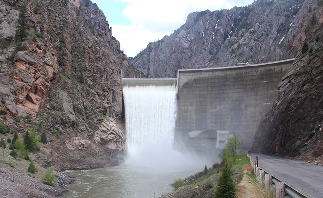 A concrete dam spilling water into a reservoir below. Steep canyon walls are around the dam structure