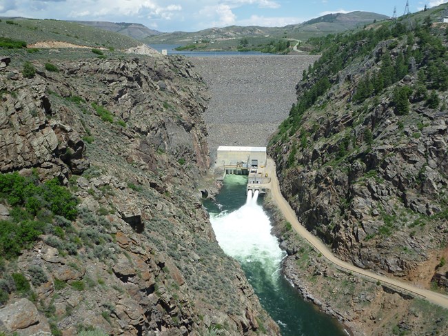 A large earth and rock dam with spillways open into a narrow reservoir