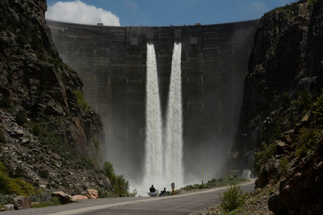 A large concrete dam with spillways open. People are standing at an overlook below it