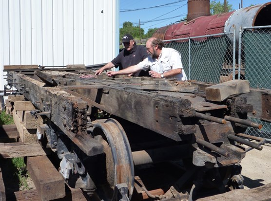 Two people look at the base of a tender rail vehicle. Rusted metal sections and wheels are visible. A white building wall and green chain link fence are behind the people.