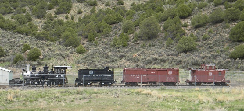 Four train cars (two black, two red) are arranged in a line. They are located in a maintenance yard with grass. Green vegetation is on a slope behind the train cars.