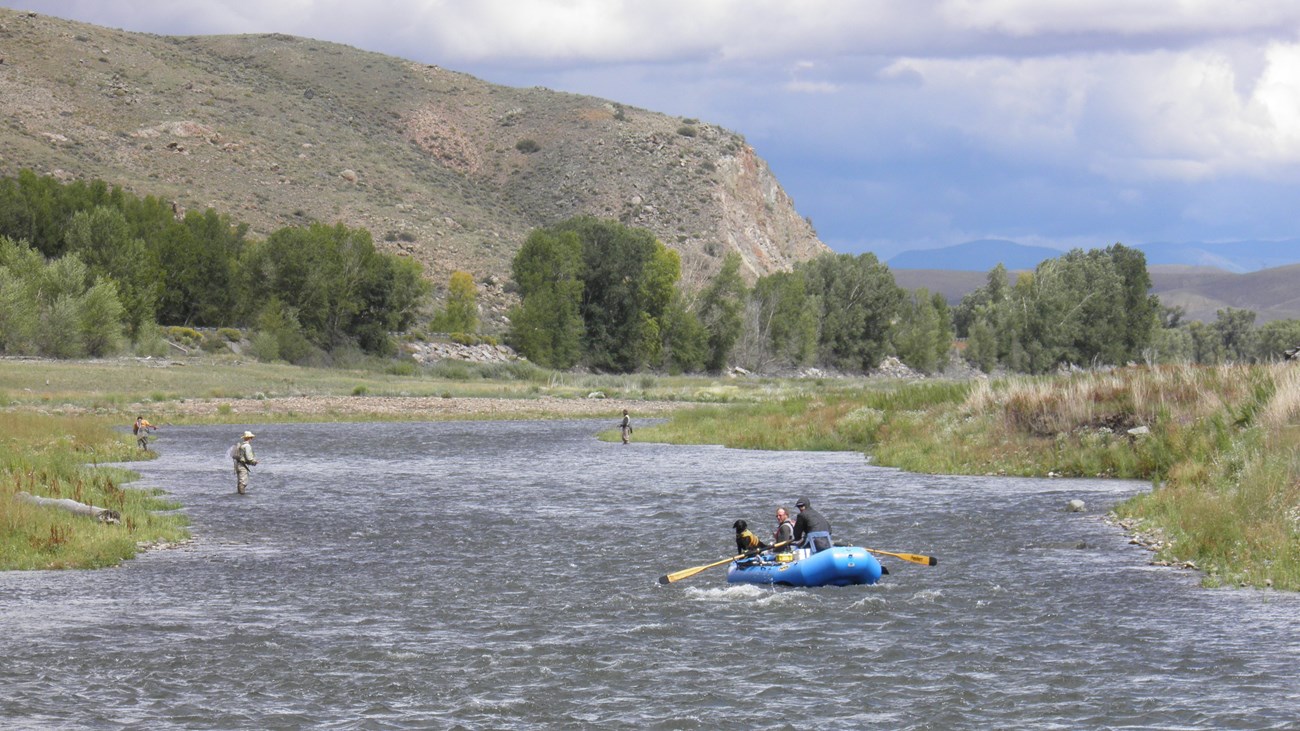 A river runs through an area with a mesa, trees, and vegetation around it. Three anglers stand in the water fishing. One blue raft with two people and a dog paddle in the foreground.