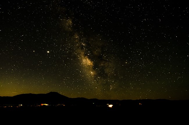 Milky Way over a faintly lit campground. Dark silhouettes of mesas are in the background.