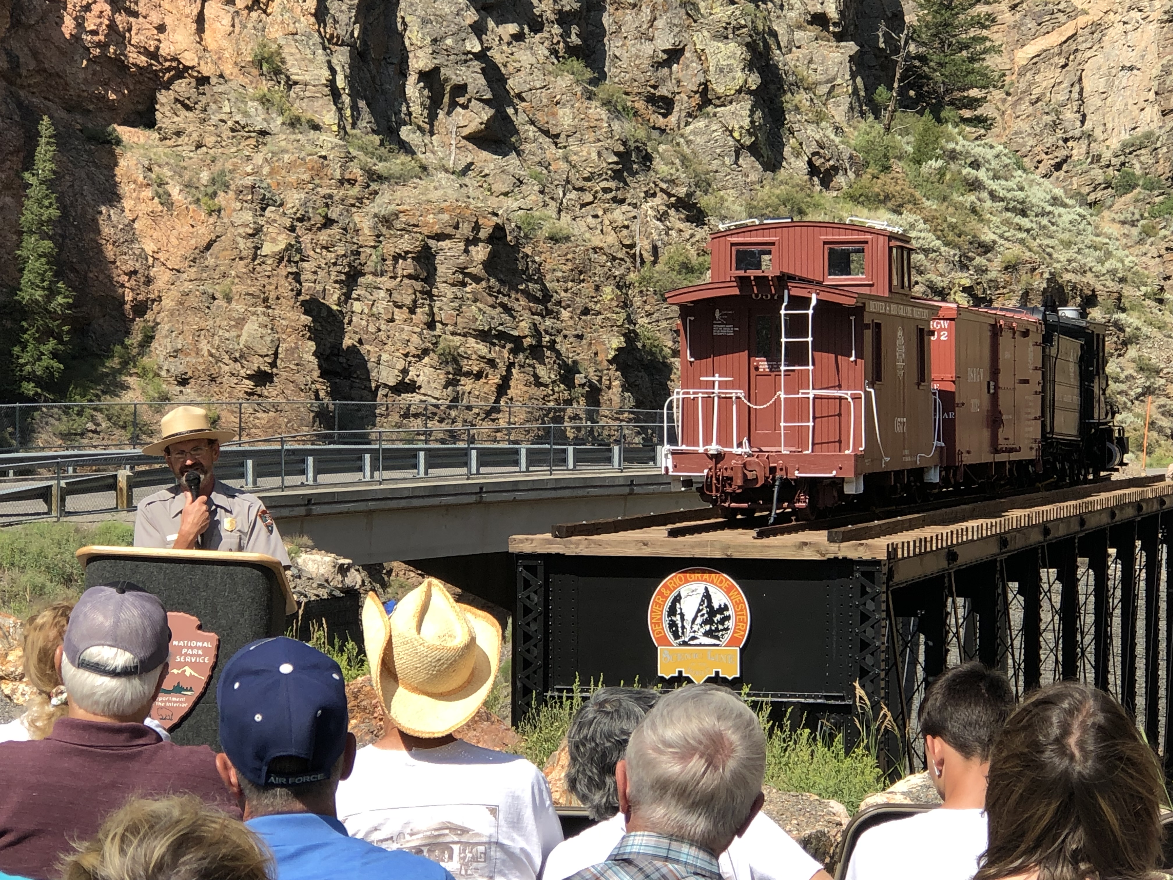 park ranger Paul Zaenger speaks at a podium in front of a narrow gauge caboose on a truss bridge in a deep, dramatic canyon