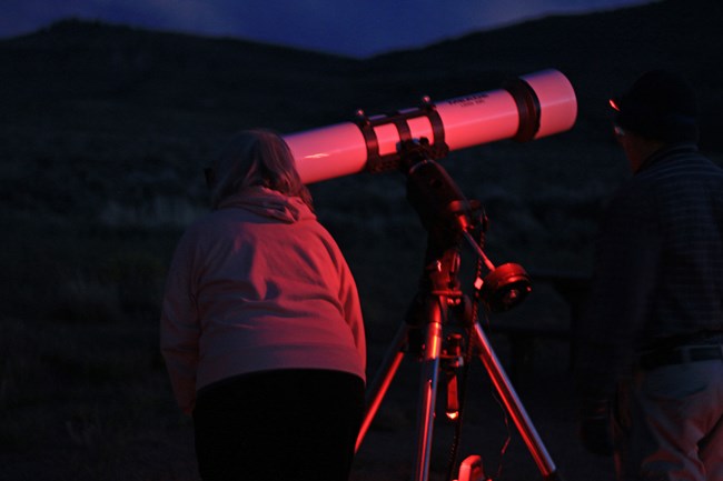 A person illuminated by red light stands by a large white telescope