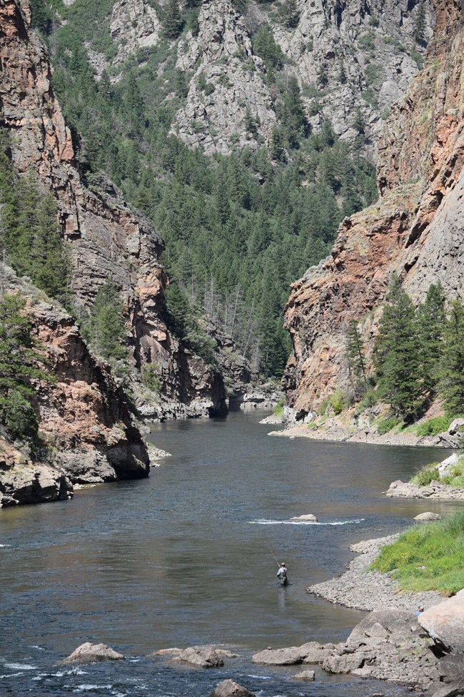 Person fly fishing in a narrow reservoir. Tall canyon walls with conifers surround the water.